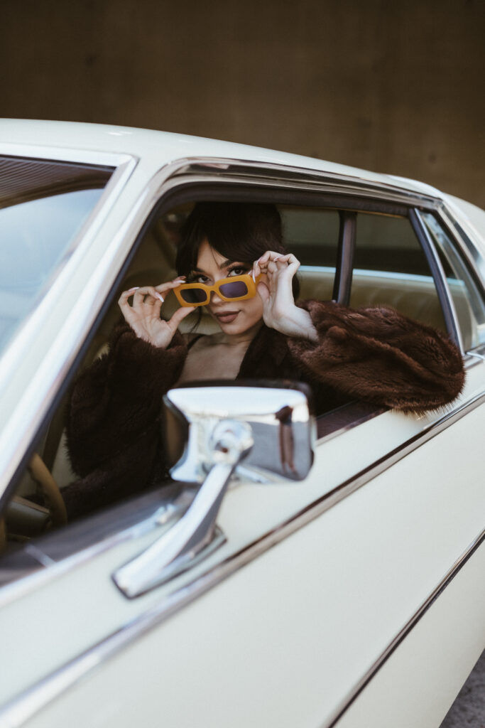 Chic woman in black lace boudoir set and fur coat, wearing sunglasses while posing inside a classic car in Portland, Oregon, radiating confidence and style.